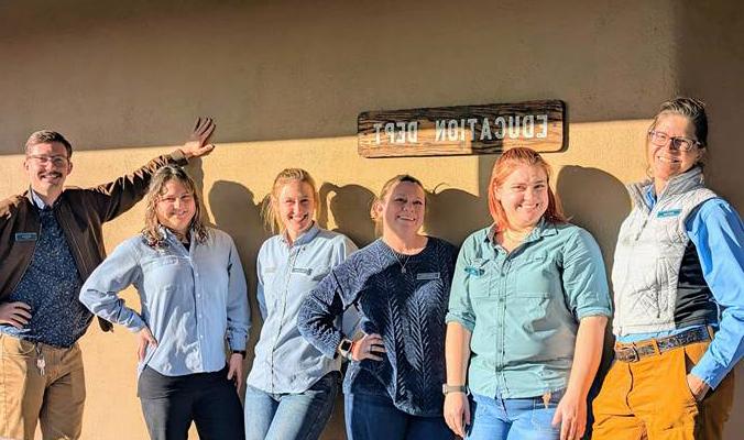 A group of six Day Camp Teachers standing in front of an adobe wall. The sun is shining and they are all smiling broadly.